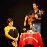 Sur un fond noir, une femme est assise à côté d'un table tonde, à sa gauche un homme debout en train de jouer une guitare